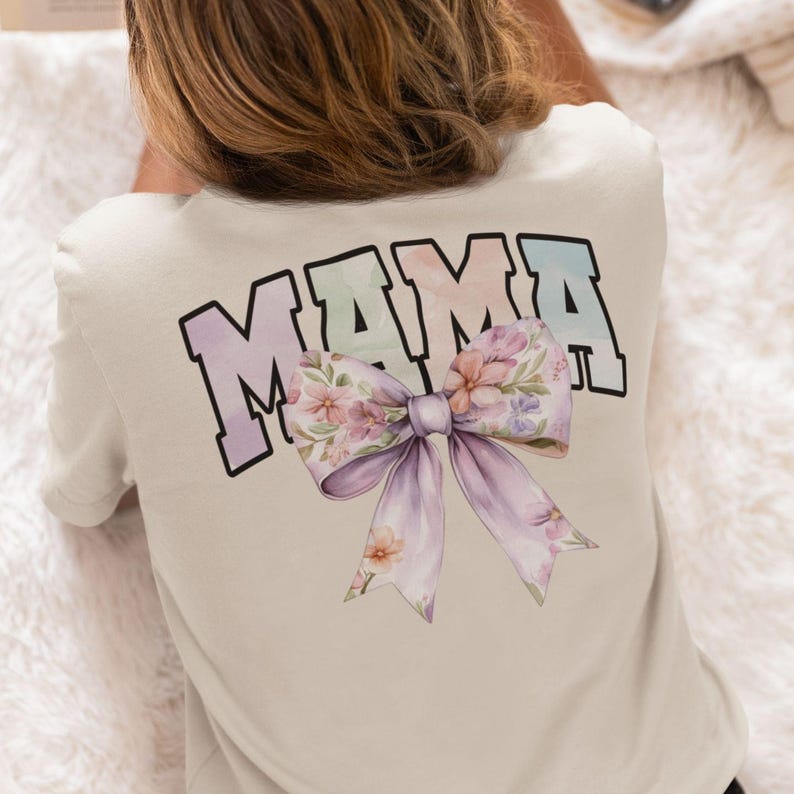 Peut inclure: Un t-shirt blanc avec le mot "MAMA" imprim&eacute; en couleurs arc-en-ciel avec un n&oelig;ud floral en dessous.