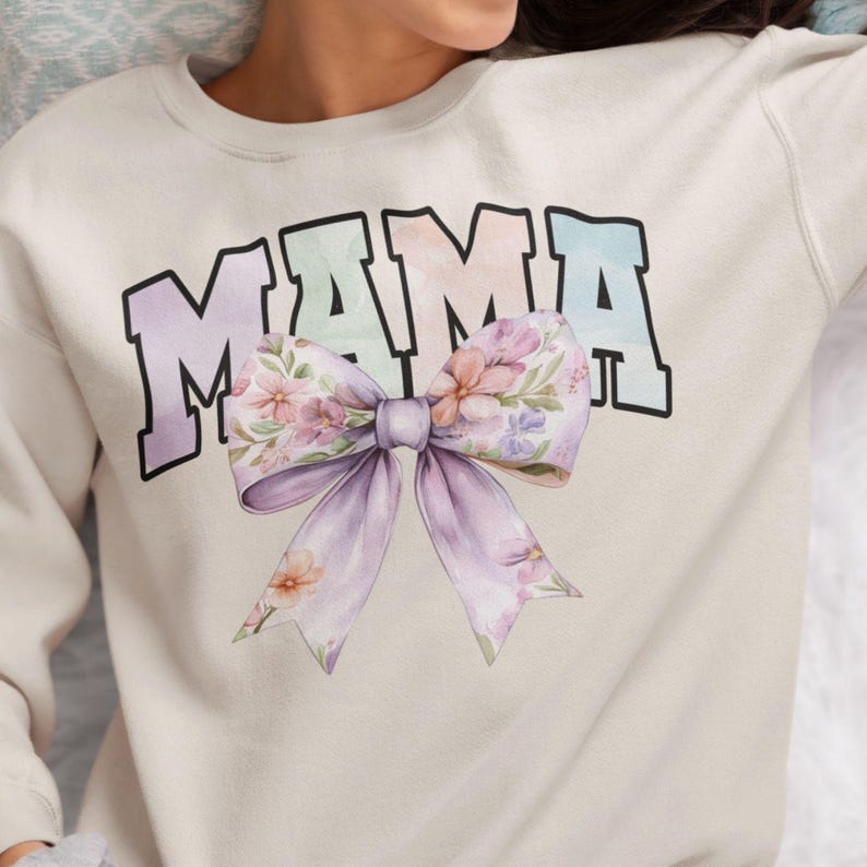 Peut inclure: Un sweat-shirt beige clair avec le mot "MAMA" imprim&eacute; en couleurs arc-en-ciel. Un grand n&oelig;ud floral est centr&eacute; sous le mot.