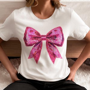 May include: White t-shirt featuring a large, sparkly pink bow graphic. The bow has a glittery texture and is centered on the shirt. The shirt has a crew neck and short sleeves.
