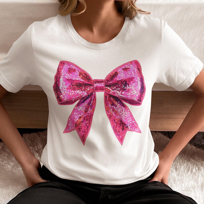 May include: White t-shirt featuring a large, sparkly pink bow graphic. The bow has a glittery texture and is centered on the shirt. The shirt has a crew neck and short sleeves.