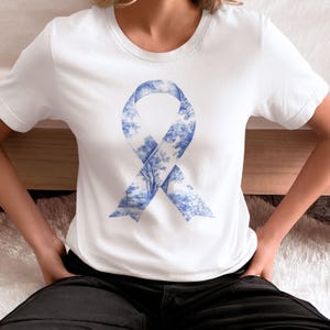 May include: White t-shirt with a blue ribbon design. The ribbon is made up of a blue and white pattern of trees and foliage.