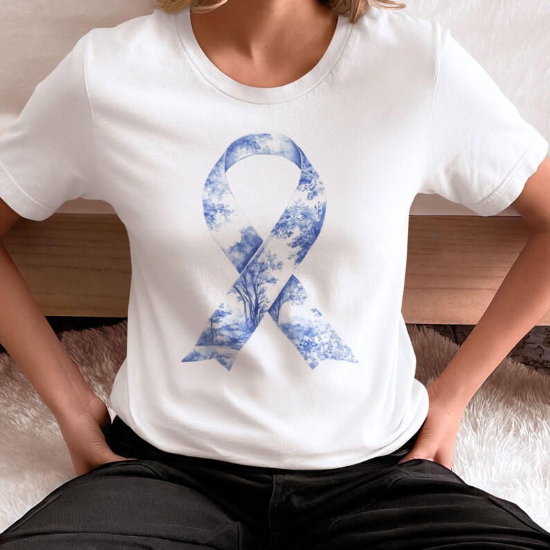 May include: White t-shirt with a blue ribbon design. The ribbon is made up of a blue and white pattern of trees and foliage.