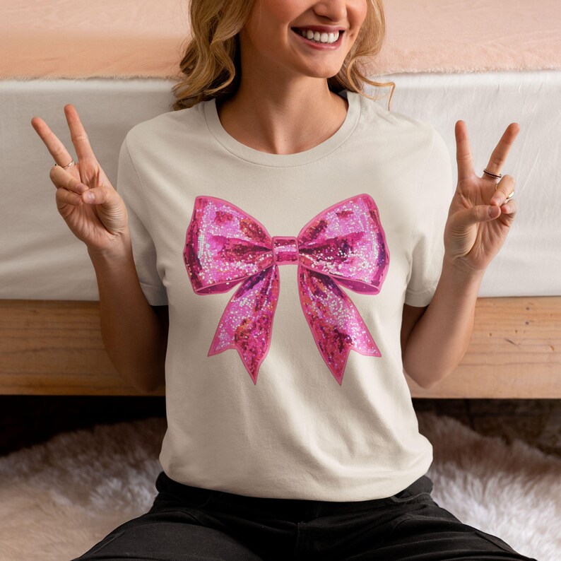 May include: A cream-colored t-shirt featuring a large, sparkly pink bow graphic. The bow has a glittery texture and is centered on the shirt. The shirt is a casual, everyday wear item.