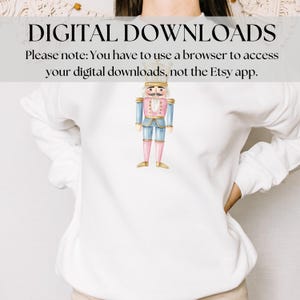 May include: White sweatshirt featuring a watercolor illustration of a nutcracker soldier. The nutcracker is dressed in a blue jacket, pink pants, and gold boots. Text at the top reads "DIGITAL DOWNLOADS".