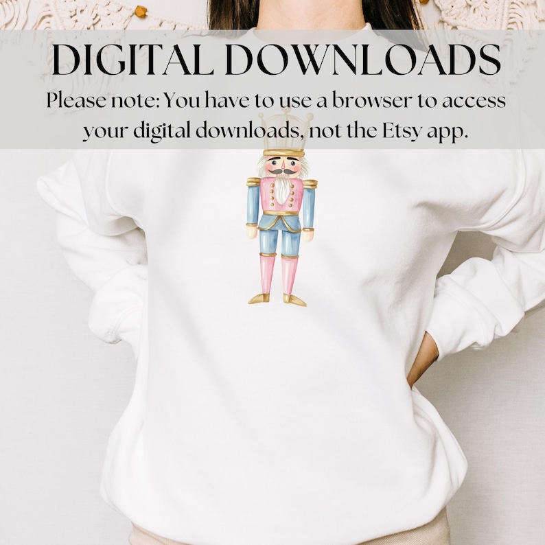 May include: White sweatshirt featuring a watercolor illustration of a nutcracker soldier. The nutcracker is dressed in a blue jacket, pink pants, and gold boots. Text at the top reads "DIGITAL DOWNLOADS".