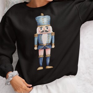 May include: Black sweatshirt featuring a watercolor illustration of a blue and gold nutcracker. The nutcracker has a white beard and mustache, and is wearing a tall hat with stars. The design is centered on the front of the sweatshirt.