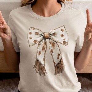 Puede incluir: Una camiseta beige con un lazo marr&oacute;n y blanco con detalles de flecos. El lazo tiene un patr&oacute;n de manchas.