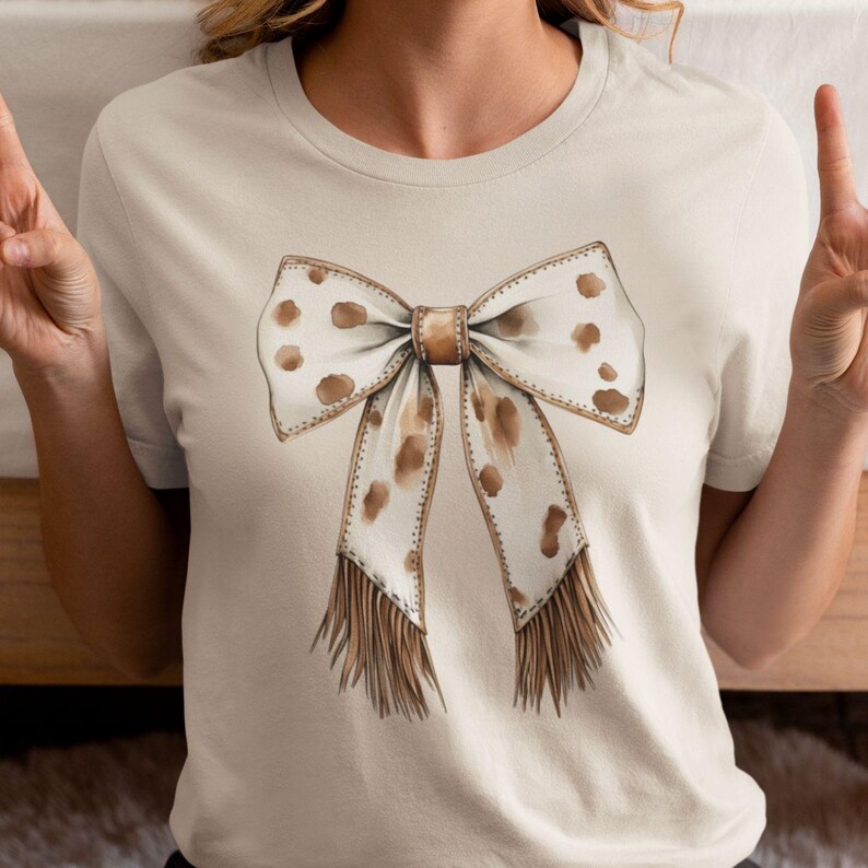 Puede incluir: Una camiseta beige con un lazo marr&oacute;n y blanco con detalles de flecos. El lazo tiene un patr&oacute;n de manchas.
