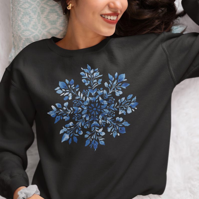 May include: A black sweatshirt with a blue watercolor snowflake design. The snowflake is made up of intricate details and has a delicate, floral appearance.