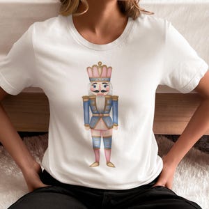May include: White t-shirt featuring a watercolor illustration of a nutcracker soldier. The nutcracker is dressed in a blue jacket, pink pants, and a pink and gold crown. The design is centered on the shirt.