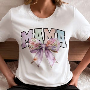 Peut inclure: T-shirt blanc avec un graphique "MAMA" en d&eacute;grad&eacute; arc-en-ciel et un n&oelig;ud floral violet.