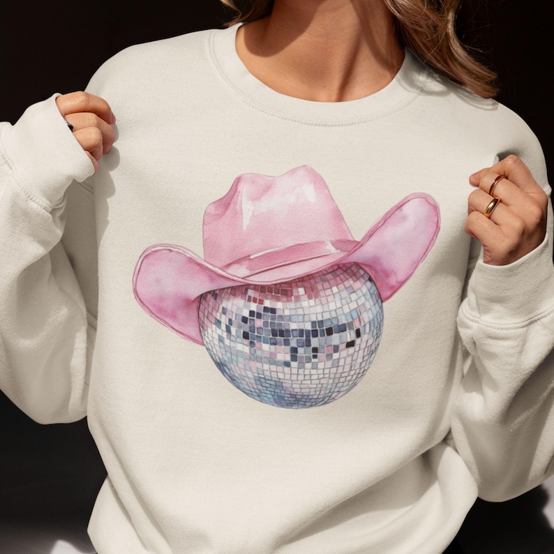 May include: A white sweatshirt with a pink cowboy hat and a silver disco ball graphic.