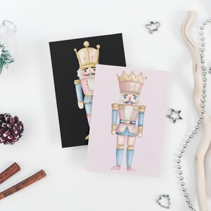 May include: Two greeting cards featuring watercolor illustrations of nutcrackers. One card is on a black background, the other on a pink background. The nutcrackers wear pink, blue, and gold uniforms and crowns. The cards are surrounded by holiday decorations.