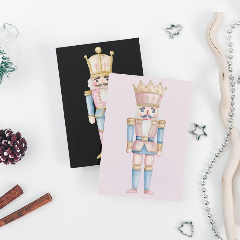May include: Two greeting cards featuring watercolor illustrations of nutcrackers. One card is on a black background, the other on a pink background. The nutcrackers wear pink, blue, and gold uniforms and crowns. The cards are surrounded by holiday decorations.
