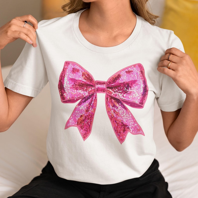 May include: White t-shirt featuring a large, sparkly pink bow graphic. The bow has a glittery texture and is centered on the front of the shirt. The shirt has a classic crew neck and short sleeves.