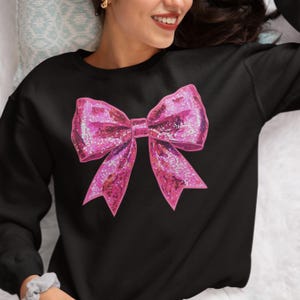 May include: Black sweatshirt featuring a large, glittery pink bow graphic. The bow has a textured appearance with sparkling accents, creating a festive and eye-catching design. The sweatshirt is a casual garment, suitable for everyday wear.