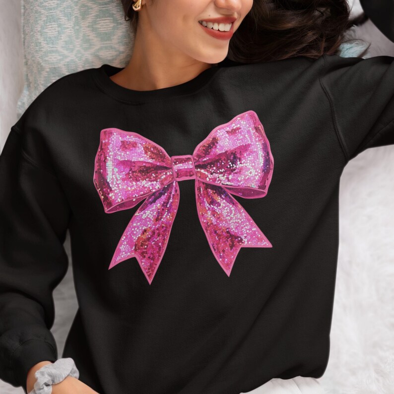 May include: Black sweatshirt featuring a large, glittery pink bow graphic. The bow has a textured appearance with sparkling accents, creating a festive and eye-catching design. The sweatshirt is a casual garment, suitable for everyday wear.