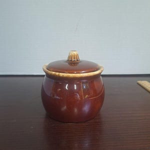 Pode incluir: Um açucareiro redondo de cerâmica castanha com tampa a condizer. A taça apresenta um esmalte castanho escuro brilhante com reflexos bege mais claros à volta do rebordo e do puxador da tampa. A tampa tem uma pequena pega estriada no centro.
