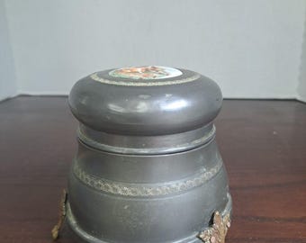 Caja de polvos musical de metal estadounidense de la década de 1930 con incrustaciones de porcelana y escena romántica.