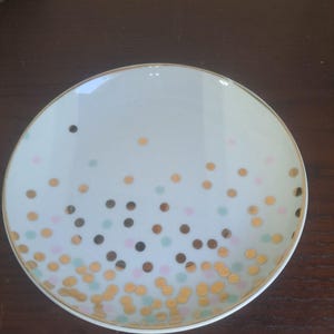 Może przedstawiać: Biały ceramiczny talerz ze złotą obwódką i wzorem konfetti z złotych, brązowych, różowych i jasnoniebieskich kropek. Talerz jest okrągły i leży na ciemnobrązowej powierzchni.