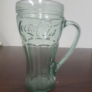 Könnte beinhalten: Ein klarer, grün getönter Glaskrug mit Henkel. Auf der Vorderseite ist das Coca-Cola-Logo eingeprägt. Der Krug hat eine einzigartige Form, die oben und unten breiter ist, mit einer schmaleren Taille.