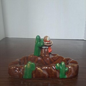 May include: A ceramic ashtray with a brown base and a sombrero-wearing figure. Green cacti are placed around the ashtray. The figure has a red scarf and a brown hat. The ashtray is on a wooden surface.