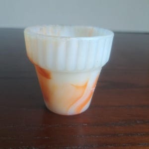 Op de afbeelding: Een kleine, doorschijnende witte glazen beker met een uitlopende, geribbelde rand en een wervelend oranje marmerpatroon over het hele lichaam. Het vat heeft een taps toelopende vorm die naar de basis toe smaller wordt, met een gladde afwerking.