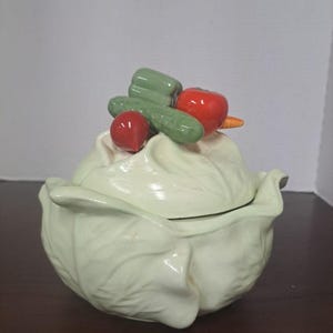 May include: A light green ceramic cabbage-shaped serving dish with a lid, adorned with ceramic vegetables. The vegetables include a red tomato, a green pepper, a cucumber, and a radish. The dish is on a dark brown surface.