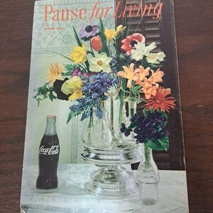 以下が含まれることがあります： 「Pause for Living」というタイトルのヴィンテージ雑誌の表紙。生け花とコカ・コーラのボトルが描かれています。花には、黄色の水仙、オレンジ色のガーベラ、紫のアネモネが含まれています。雑誌は1950年春号です。