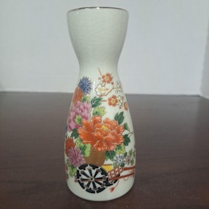 May include: A white ceramic sake bottle with a floral design. The bottle features a colorful arrangement of orange, pink, and blue flowers, along with a decorative cart. The bottle has a crackled glaze and a gold rim.