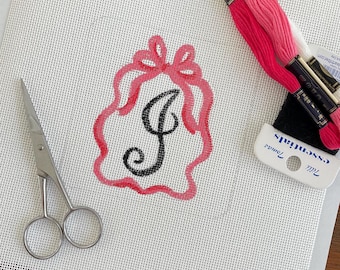 May include: A needlepoint canvas featuring a hand-painted black letter J centered within a pink scalloped border topped with a bow. Beside the canvas are silver metal embroidery scissors and two skeins of pink embroidery floss labeled Tilli Tomas essentials.