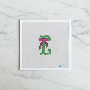May include: A square canvas with a green letter "L" and a pink bow. The letter is stitched in a cross-stitch style. The lower right corner has the text "NCC" in blue.