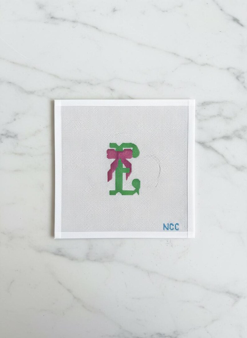 May include: A square canvas with a green letter "L" and a pink bow. The letter is stitched in a cross-stitch style. The lower right corner has the text "NCC" in blue.