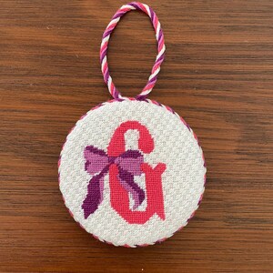 May include: A round, white cross-stitch ornament featuring a pink ampersand symbol with a purple bow. The ornament has a twisted cord loop in shades of pink, purple, and white for hanging. The edge is trimmed with a pink and purple border.