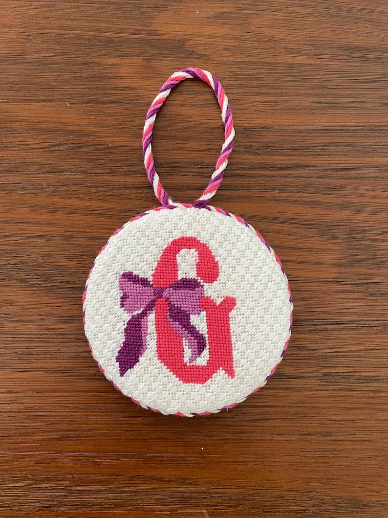 May include: A round, white cross-stitch ornament featuring a pink ampersand symbol with a purple bow. The ornament has a twisted cord loop in shades of pink, purple, and white for hanging. The edge is trimmed with a pink and purple border.