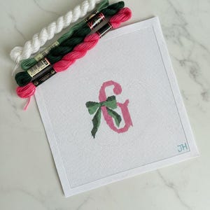May include: A white needlepoint canvas with a pink ampersand and green bow design. Strands of white, green, and pink embroidery floss are arranged above the canvas. The canvas has a stitched border and the initials "JH" in the corner.