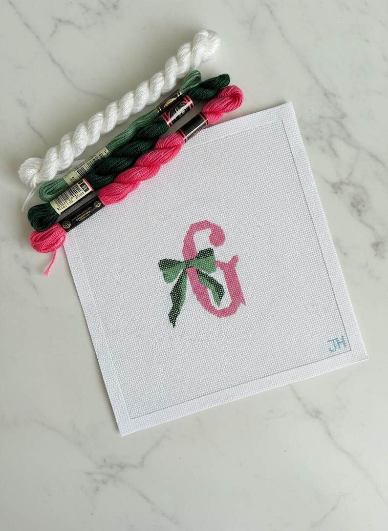 May include: A white needlepoint canvas with a pink ampersand and green bow design. Strands of white, green, and pink embroidery floss are arranged above the canvas. The canvas has a stitched border and the initials "JH" in the corner.