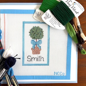 May include: A needlepoint canvas featuring a topiary design with a blue bow and the name "Smith". The canvas is framed in blue, with various spools of green, brown, and blue embroidery floss. The text "Very Velvet" is visible on the floss.