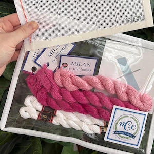 May include: A needlepoint kit featuring a white mesh canvas with a partial pink design, contained in a clear plastic pouch. Inside are skeins of pink and white embroidery thread, labels reading MILAN by tilli tomas, and a Nashville Canvas Co logo.