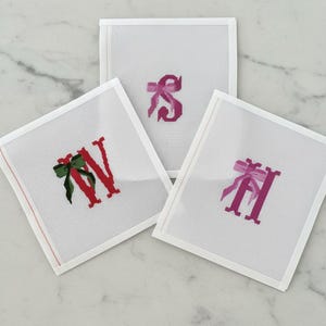 May include: Three white canvas squares with embroidered letters and bows. The letters are red and pink, with green and pink bows. The squares are arranged on a white marble surface.