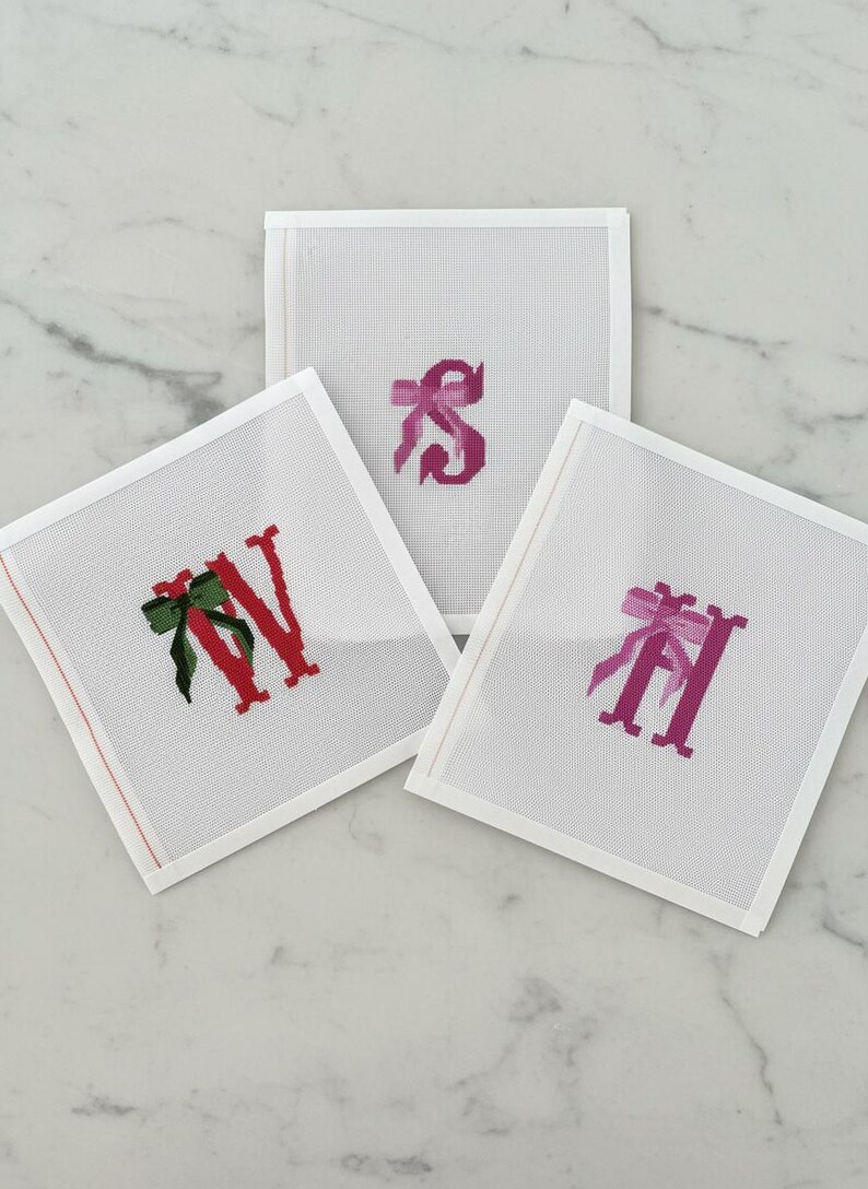 May include: Three white canvas squares with embroidered letters and bows. The letters are red and pink, with green and pink bows. The squares are arranged on a white marble surface.