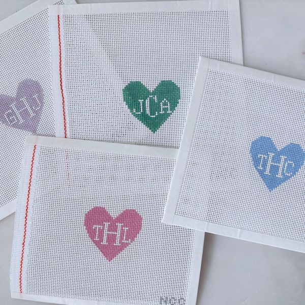 Può includere: Quattro tele rettangolari bianche per ricamo con disegni di cuori centrati in lavanda, verde, blu e rosa. Ogni cuore contiene un monogramma di tre lettere ricamato in bianco. Le tele presentano una linea di bordo verticale rossa.