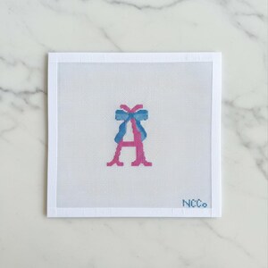 May include: A needlepoint canvas featuring a pink letter "A" with a blue bow at the top. The canvas is white with a grid pattern, and the text "NCCo." is in the bottom right corner. The background is a marble surface.