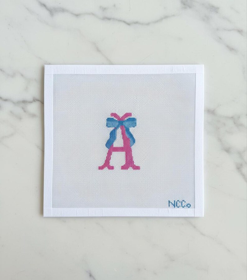 May include: A needlepoint canvas featuring a pink letter "A" with a blue bow at the top. The canvas is white with a grid pattern, and the text "NCCo." is in the bottom right corner. The background is a marble surface.