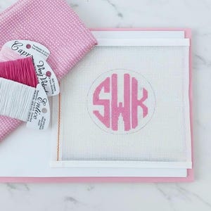 May include: A pink needlepoint kit featuring a monogram with the letters SWK in a circle. The kit includes pink and white embroidery floss, a pink polka dot fabric, and a white canvas. The kit is designed for embroidery and needlepoint projects.