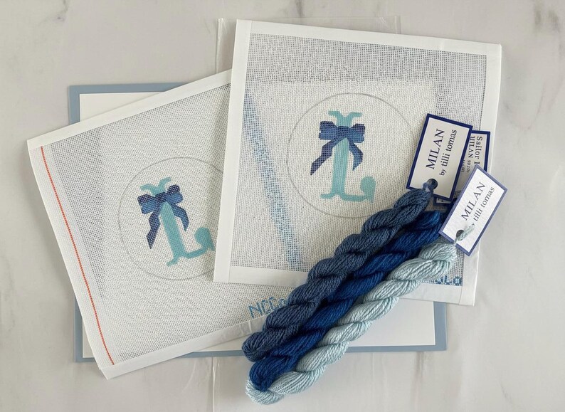 May include: Needlepoint kit featuring a monogrammed "L" with a bow in shades of blue. The kit includes canvas, thread, and tags. The canvas is white with a blue design. The thread is in various shades of blue.
