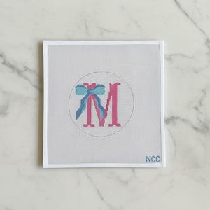 May include: A needlepoint canvas featuring a pink letter "M" with a blue bow design inside a white circle. The canvas is on a white background with the letters "NCC" in the bottom right corner.