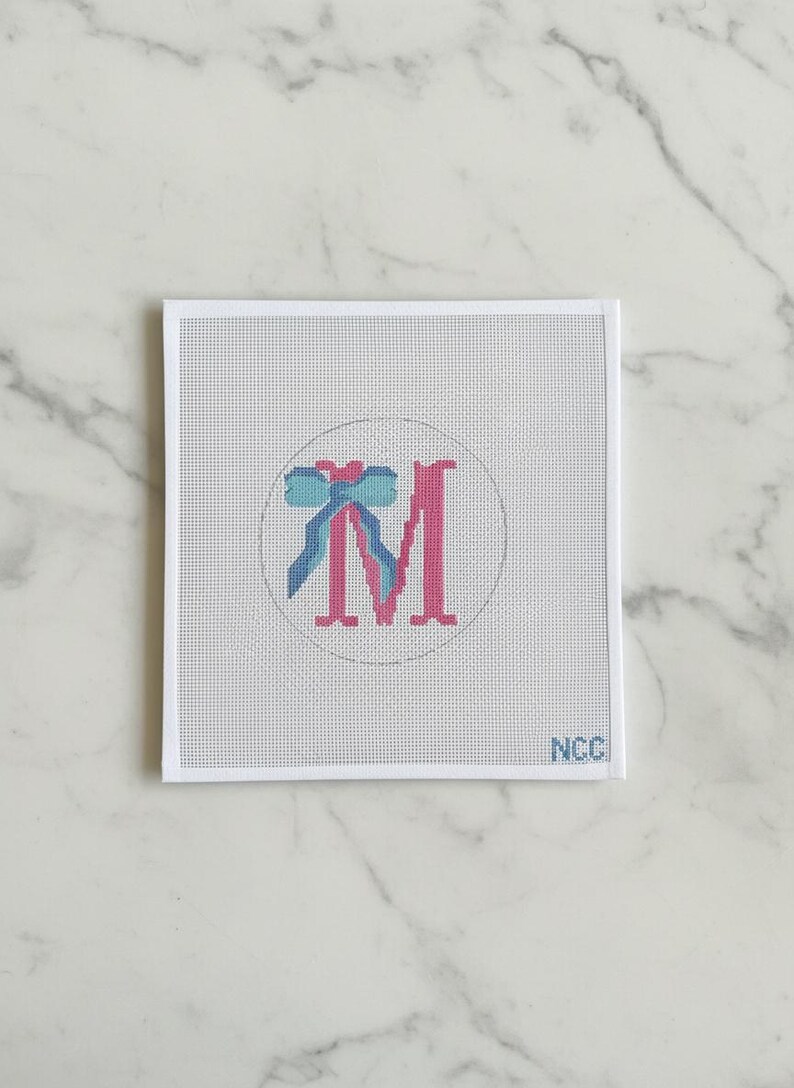 May include: A needlepoint canvas featuring a pink letter "M" with a blue bow design inside a white circle. The canvas is on a white background with the letters "NCC" in the bottom right corner.