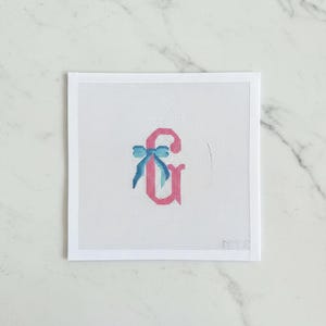 May include: Needlepoint canvas featuring a pink ampersand with a blue bow. The ampersand is stitched on a white mesh canvas, set against a white background. The design is simple and elegant, suitable for personalized crafts.