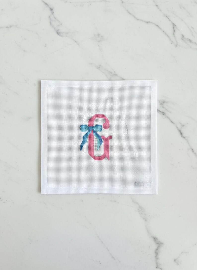 May include: Needlepoint canvas featuring a pink ampersand with a blue bow. The ampersand is stitched on a white mesh canvas, set against a white background. The design is simple and elegant, suitable for personalized crafts.